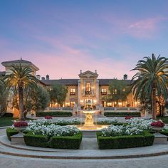 a large mansion with a fountain surrounded by flowers and trees in front of it at dusk