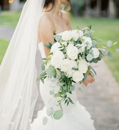 a bride holding a bouquet of white roses and greenery in her hand while walking down a path