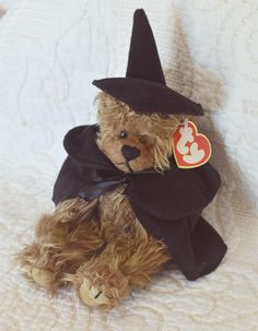 a teddy bear dressed up as a witch sitting on a white bed with a heart tag