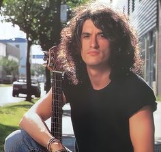 a man with long hair sitting on the ground holding an acoustic guitar in his hands