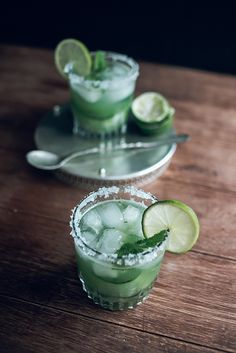 two glasses filled with green drinks on top of a wooden table