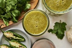 two glasses filled with green smoothie next to some fresh vegetables on a white table