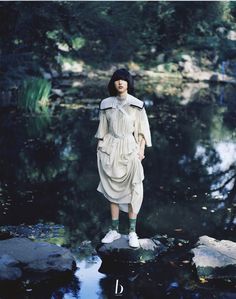 a woman standing on top of a rock next to a body of water with trees in the background