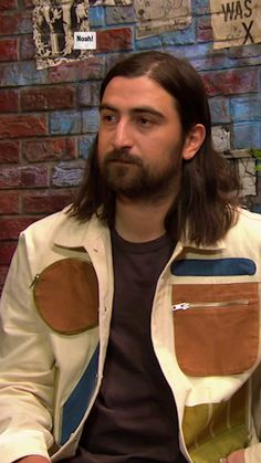 a man with long hair sitting in front of a brick wall wearing a white jacket