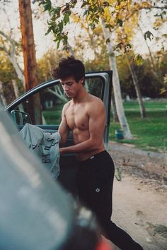 a shirtless young man standing next to a car with his hand on the door handle