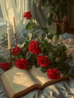 an open book sitting on top of a bed next to red roses and two candles