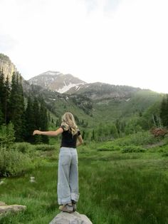 a woman standing on top of a rock in the middle of a field with mountains behind her