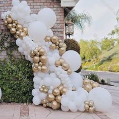 white and gold balloons in front of a brick wall with bushes behind it on the sidewalk