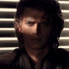 a man with long hair and blue eyes wearing a black shirt in front of blinds