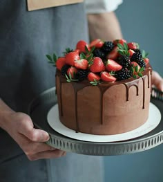 a person holding a chocolate cake with strawberries and blackberries on top