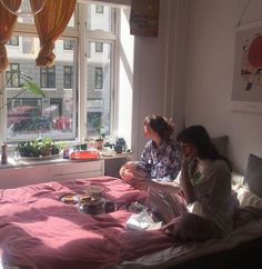 two people are sitting on a bed in front of a window and eating food together