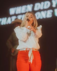 a woman in red pants and white shirt singing into a microphone with words projected on the wall behind her