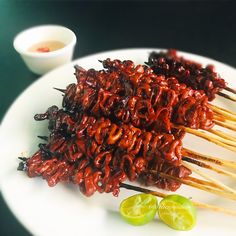 several skewers of food on a white plate with limes and dipping sauce