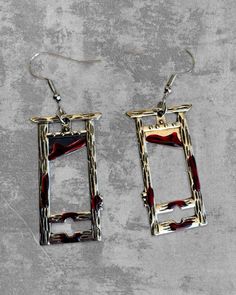 two square shaped earrings with chains hanging from them on a concrete surface, one is red and the other is gold