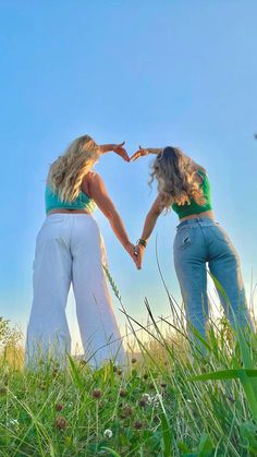 two women standing in the grass holding hands and making a heart shape with their hands