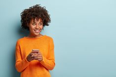 a woman in an orange sweater is looking at her cell phone and smiling while standing against a blue wall