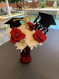 red and white flowers in a vase with graduation caps on top at a table near a pool