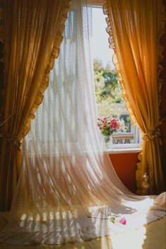 an open window with sheer curtains and flowers on the floor in front of it, next to a bed