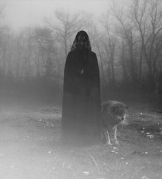 a person in a black cloak standing next to a dog on a foggy field