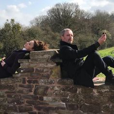 two people are sitting on a brick wall and one is taking a selfie with his cell phone