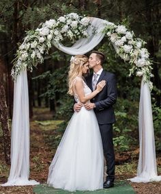 a bride and groom standing under an arch decorated with white flowers, greenery and tulle