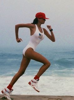 a woman running on the beach in a white swimsuit and red hat with her arm raised