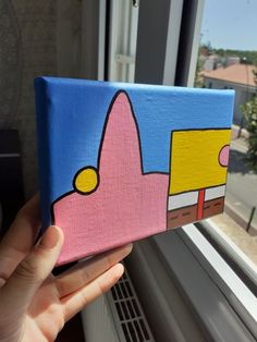 a hand holding up a small piece of art on a window sill near a building