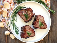 three pieces of meat on a white plate with rosemary sprigs and garlic next to it