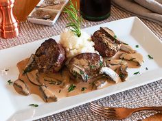 a white plate topped with meat covered in gravy and mushrooms next to mashed potatoes