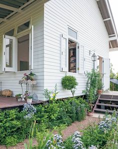 a white house with lots of plants and flowers around it's front porch area