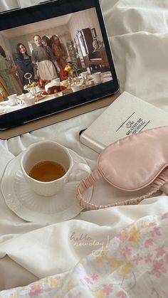 a tea cup and book on a bed next to a digital tablet with an image of women in the background