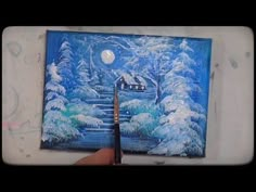 someone is holding a paintbrush in front of a snowy scene with a house and trees