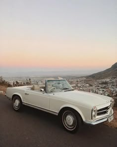 a white convertible parked on the side of a road next to a hill with a city in the background