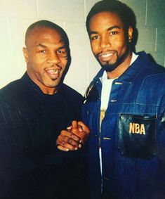 two men standing next to each other in front of a white wall and one is wearing a jacket