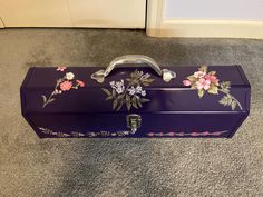 a purple suitcase with flowers painted on the lid and handles sitting on carpeted floor