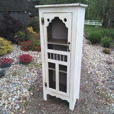 a tall white cabinet sitting on top of a gravel covered ground next to bushes and flowers