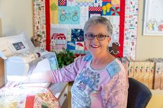 an older woman sitting at a sewing machine in front of a quilted wall hanging