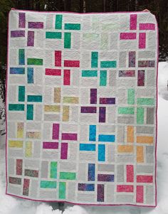 a multicolored quilt hanging from a tree in the snow with trees in the background