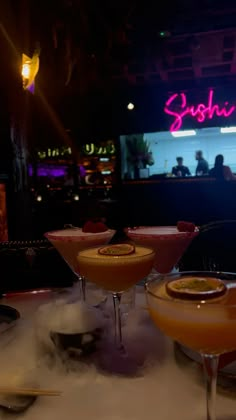 three cocktails sitting on top of a table in front of a neon sign that reads sushi