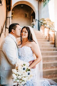 a bride and groom kissing in front of the stairs at their wedding reception, with flowers on the bouquet