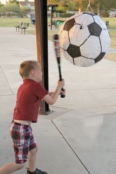 a young boy holding a baseball bat and hitting a soccer ball on a pole outside