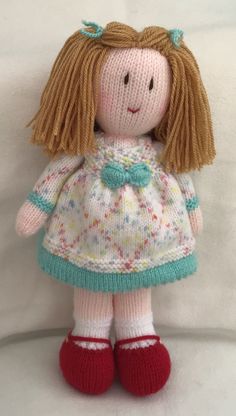 a knitted doll with blonde hair wearing a white dress and red shoes, sitting on a white surface