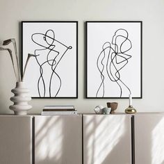 two black and white art prints on a wall above a cabinet with books, vases and lamp