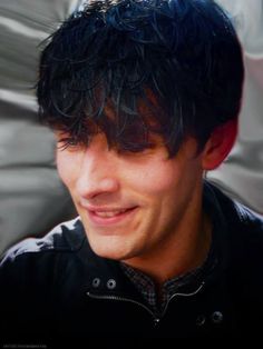 a close up of a person wearing a black jacket and smiling at the camera with hair blowing in the wind