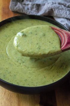 a wooden spoon full of green sauce on top of a black bowl filled with broccoli