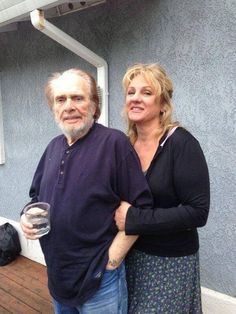 a man and woman standing next to each other on a wooden deck with a blue building in the background