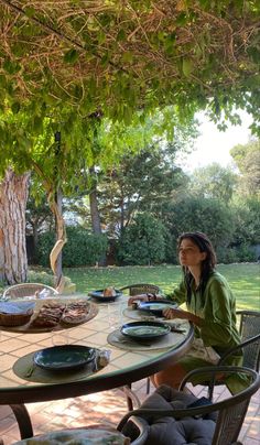 a woman sitting at a table with plates and bowls on it in front of a tree