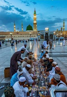 many people are sitting at a long table in the middle of an open area with lights on