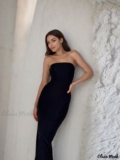 a woman in a black dress leaning against a white wall with her hands on her hips