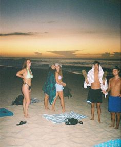 four people standing on the beach at sunset with towels and hats around their necks, one person holding a surfboard